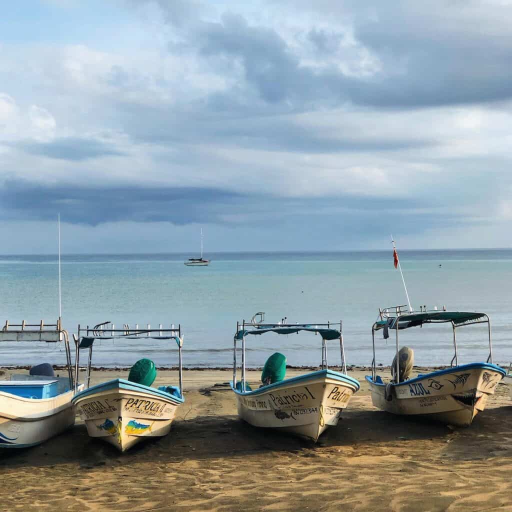 Small fishing boats lined up on the beach at Little Corn Island, Nicaragua, with calm turquoise waters and a sailboat in the distance.