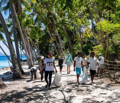 Travelers walking along a shaded beach path, carrying trash bags and picking up litter.