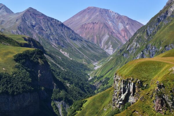 Panoramic view of the Caucasus Mountains in Georgia, showcasing steep rocky cliffs adorned with lush green hills and forests, rising dramatically against a clear blue sky with scattered clouds.