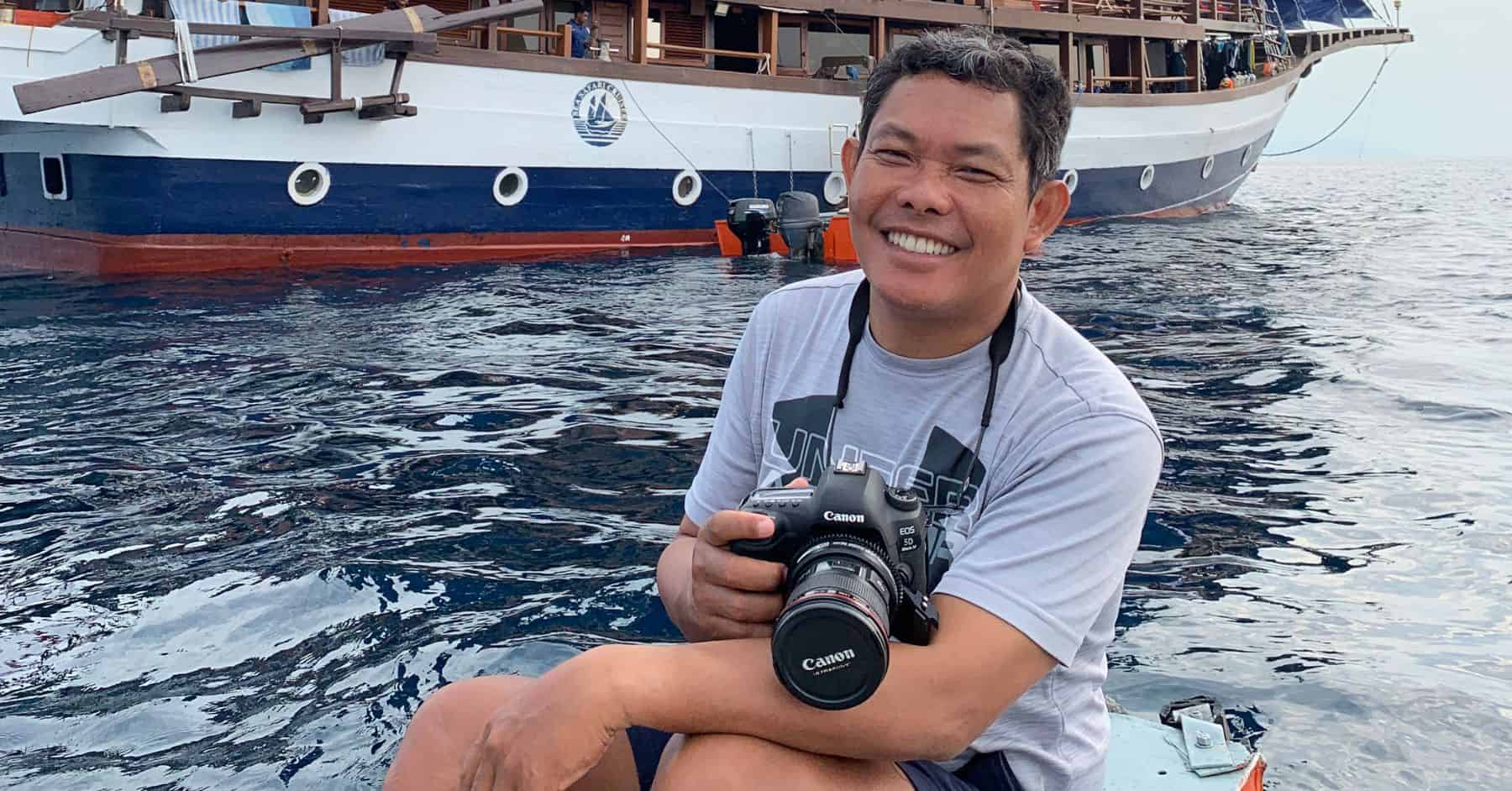 Field guide sits on dingy holding Canon DSLR camera in front of rraditional pinisi schooner boat in background. 