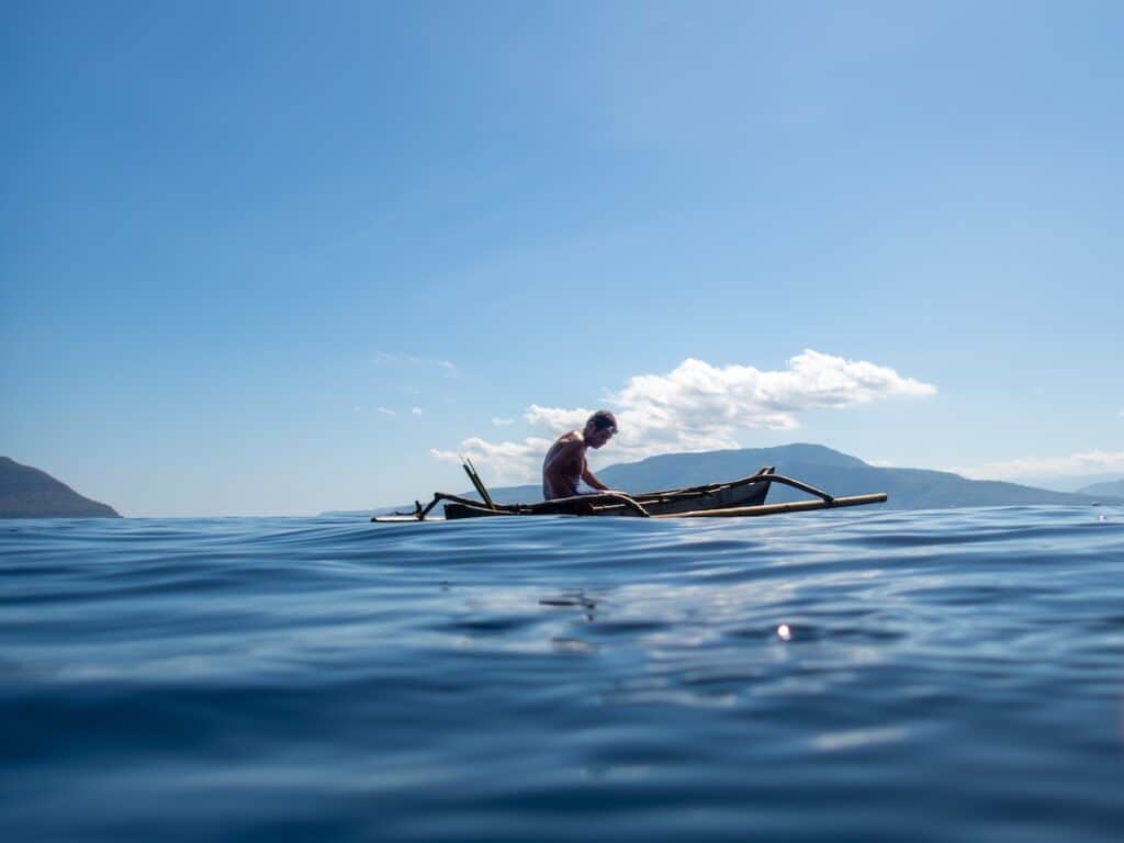 A shirtless Indonesian man with swim goggles on his head, wearing blue swim shorts, paddles a traditional bamboo outrigger canoe across calm turquoise ocean waters under a clear blue sky. Distant green volcanic mountains rise on the horizon, with scattered white clouds above.