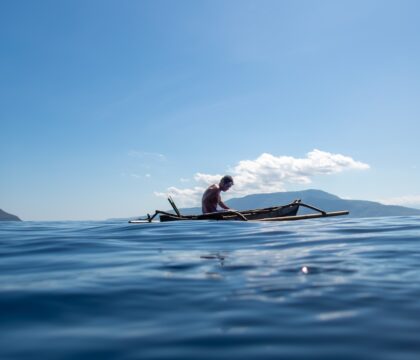 A shirtless Indonesian man with swim goggles on his head, wearing blue swim shorts, paddles a traditional bamboo outrigger canoe across calm turquoise ocean waters under a clear blue sky. Distant green volcanic mountains rise on the horizon, with scattered white clouds above.