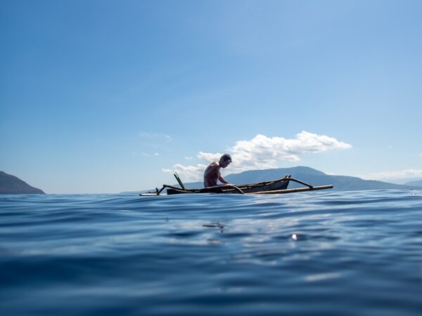 A shirtless Indonesian man with swim goggles on his head, wearing blue swim shorts, paddles a traditional bamboo outrigger canoe across calm turquoise ocean waters under a clear blue sky. Distant green volcanic mountains rise on the horizon, with scattered white clouds above.