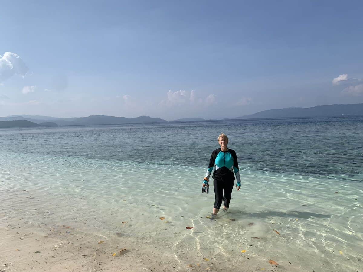 Woman standing in ankle height clear blue water holding underwater camera before entering water to snorkel