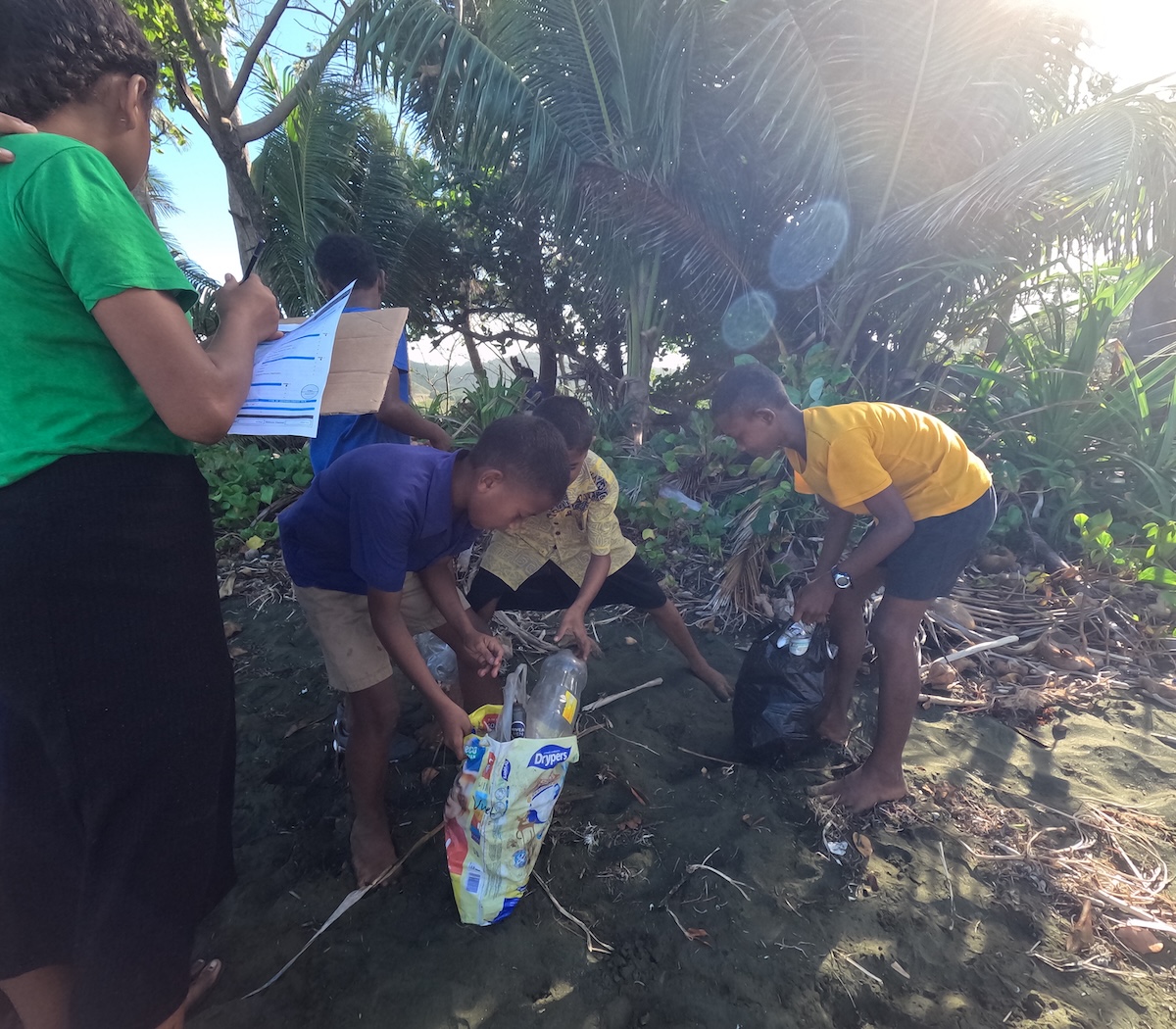A group of Fijian children sort collected plastic bottles and other waste into bags while one child records data on a clipboard during a community-led marine pollution cleanup to reduce threats to nearby coral reefs.