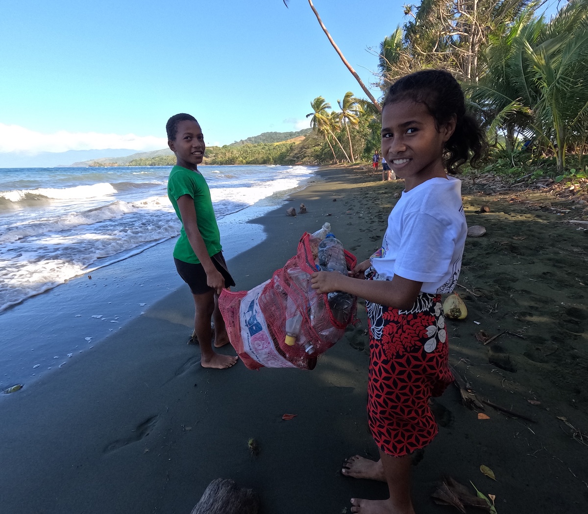 Two smiling Fijian children stand on a black sand beach at sunset with ocean waves behind them; the boy in a green shirt and the girl in a white top and red patterned skirt hold a large red mesh bag filled with collected plastic bottles and other marine debris from a community cleanup supporting reef health.