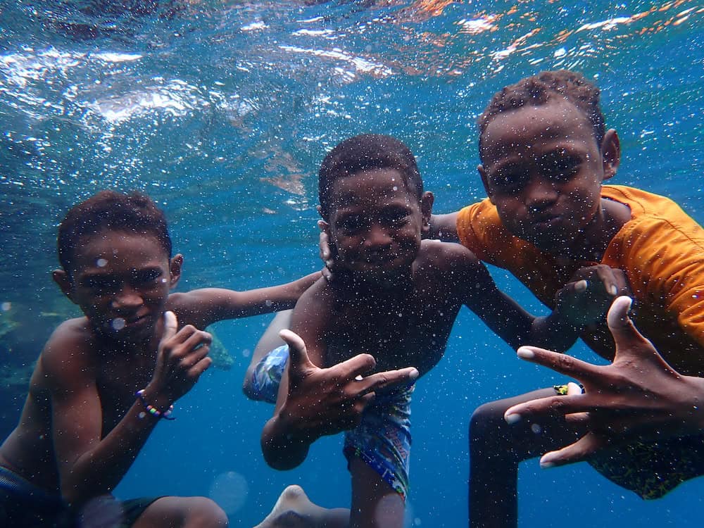 Three smiling local boys from Penemu posing playfully underwater during a spontaneous, laughter-filled photo session at the end of our morning snorkel along the south side of Penemu, Raja Ampat.