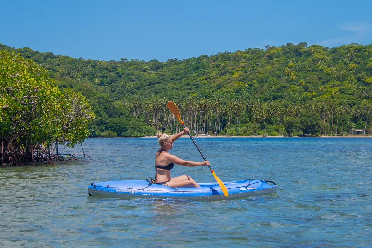 A woman with blonde hair in a black bikini top paddles a blue inflatable kayak in the calm turquoise waters of Sau Bay, Fiji.