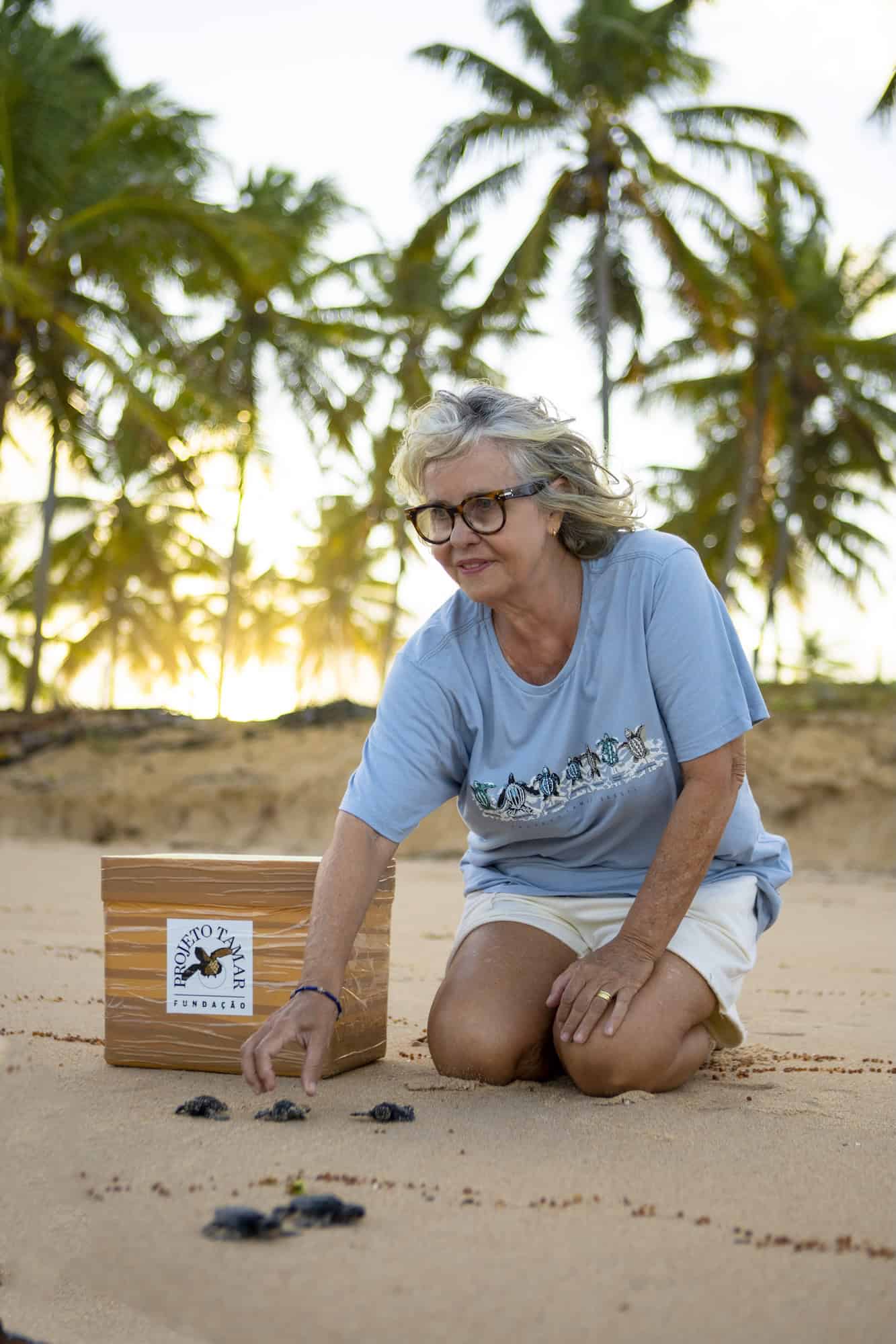 Neca Azevedo Marcovaldi, who leads Brazil’s National Sea Turtle Conservation Program known as TAMAR, kneels smiling on a sandy beach beside newly hatched sea turtle hatchlings, with palm trees in the background.