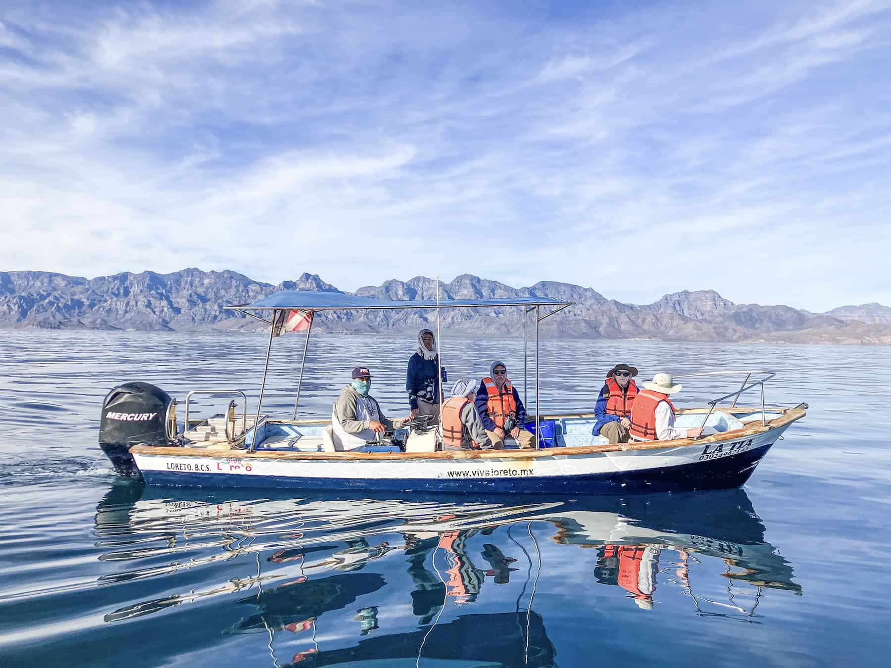 Passengers and crew aboard the panga La Tia on a mirror-calm Sea of Cortez, with the rugged mountains of Baja California reflected in the water.