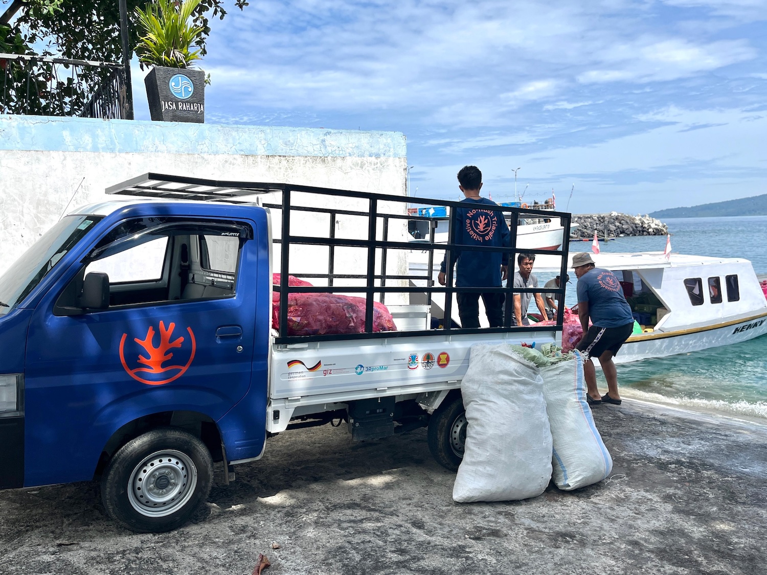 An NTTI-branded blue pickup truck is loaded with large bags of plastic waste at a harbor, with a boat docked alongside and the ocean visible in the background.