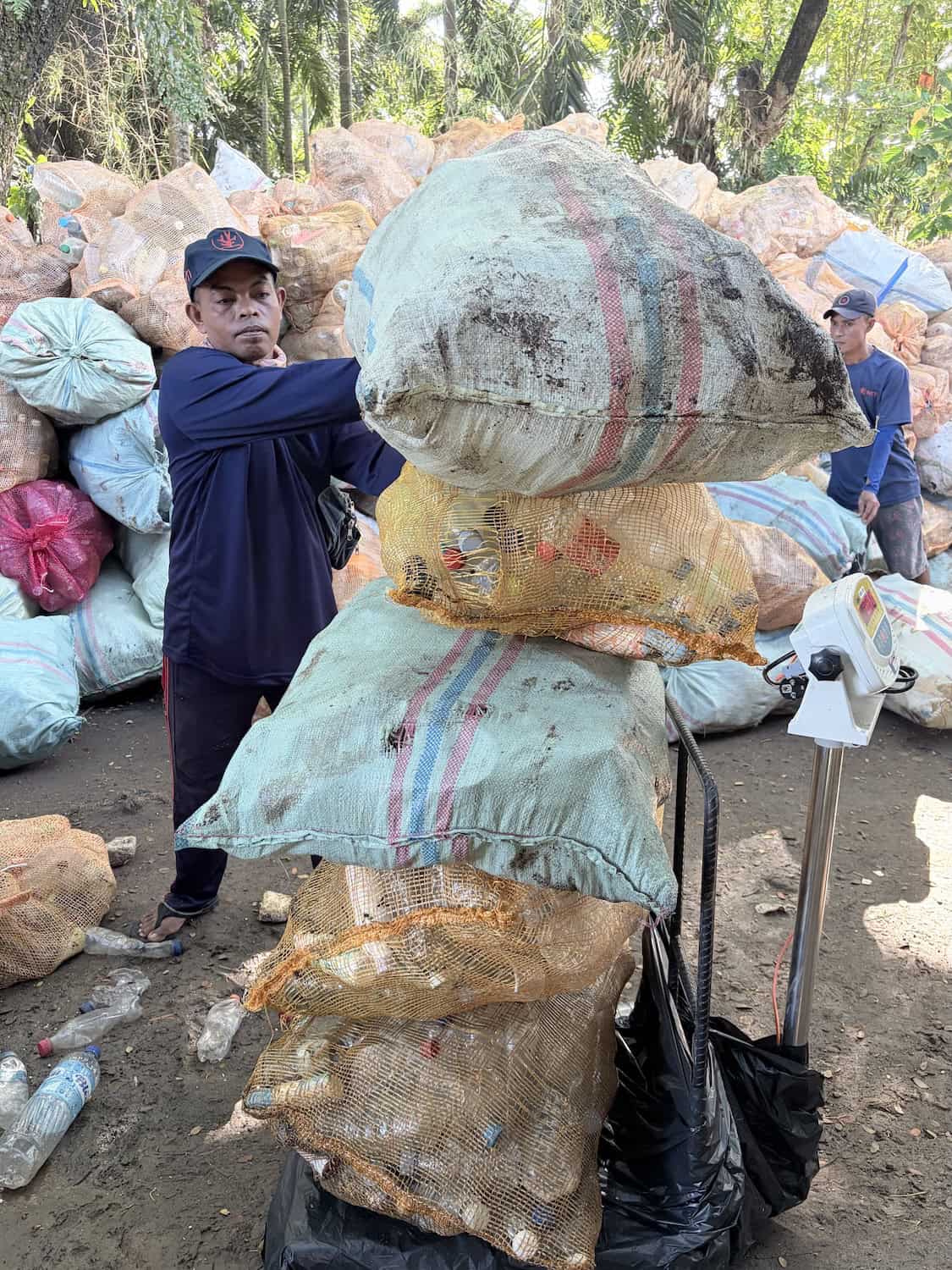 NTTI team members in navy uniforms stand among a large pile of bulging orange mesh bags filled with plastic waste, surrounded by dense tropical vegetation.