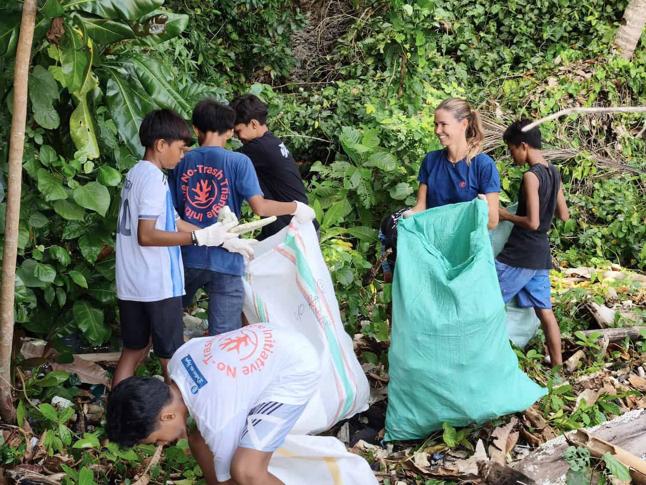 A smiling female volunteer in an NTTI shirt holds open a large green bag while local children and youth in NTTI shirts collect and deposit plastic waste along a debris-strewn jungle edge.