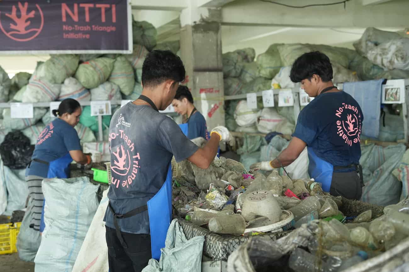 Three No Trash Triangle Initiative workers in blue uniforms sort through a large pile of plastic waste on a net-covered table inside a sorting facility, with dozens of filled bags stacked behind them.