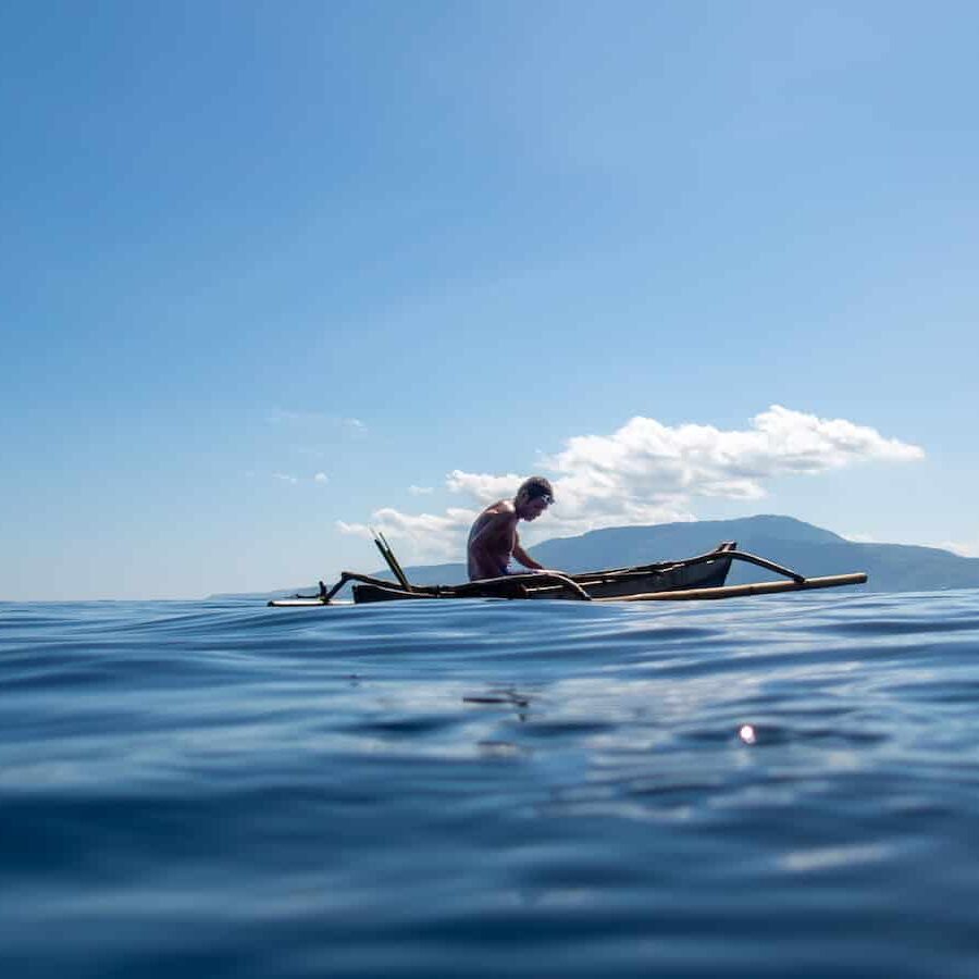 A shirtless Indonesian man with swim goggles on his head, wearing blue swim shorts, paddles a traditional bamboo outrigger canoe across calm turquoise ocean waters under a clear blue sky. Distant green volcanic mountains rise on the horizon, with scattered white clouds above.