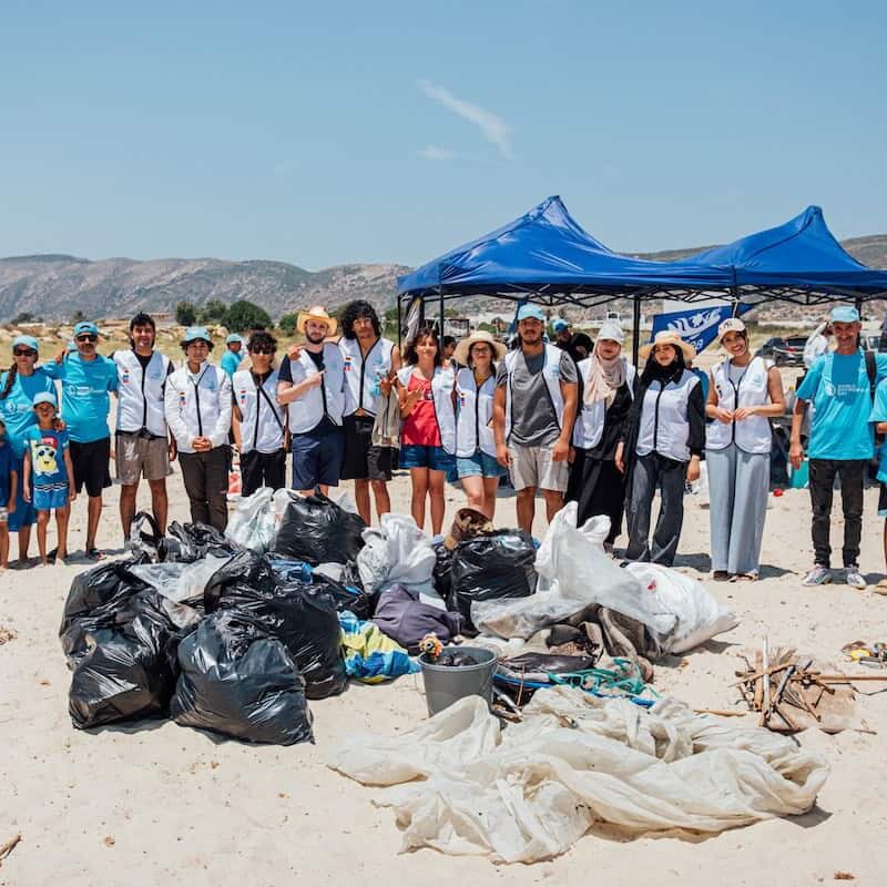 A large group of volunteers from the TunSea Organization stands on a sandy beach in Tunisia, smiling in front of piles of collected trash, including plastic bags, nets, and metal cans, after a cleanup event. Blue tents and mountains are visible in the background.